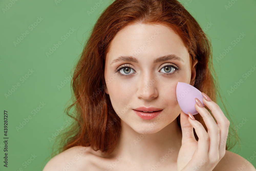 Beautiful nice half naked topless redhead hair woman 20s do nude make up apply foundation by sponge on face isolated on pastel green color background Skin care healthcare cosmetic procedures concept. Beautiful nice half naked topless redhead hair woman 20s do nude make up apply foundation by sponge on face isolated on pastel green color background Skin care healthcare cosmetic procedures concept.