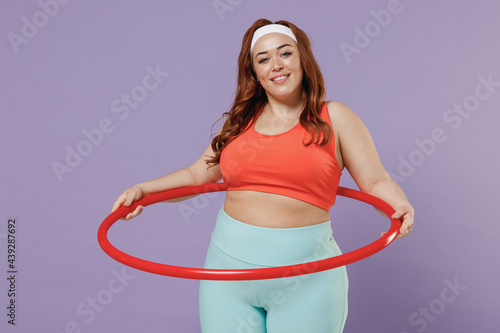 Young smiling satisfied fun chubby overweight plus size big fat fit woman 20s wearing red top warm up training twist hula hoop isolated on purple background gym home. Workout motivation sport concept