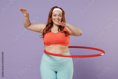 Young excited happy fun chubby overweight plus size big fat fit woman 20s wearing red top warm up training twist hula hoop isolated on purple background gym home. Workout motivation sport concept