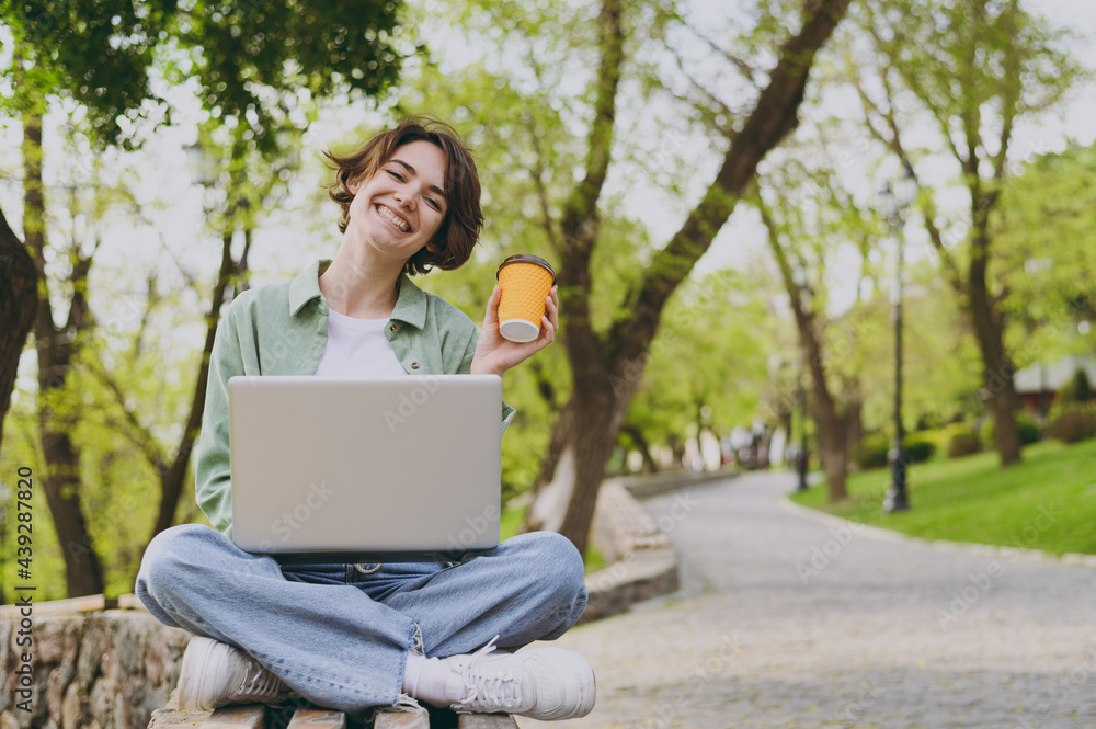 Young fun freelancer woman in green jacket jeans sit on bench in park outdoors use laptop pc computer hold takeaway delivery craft paper brown cup drink coffee to go People urban lifestyle concept Young fun freelancer woman in green jacket jeans sit on bench in park outdoors use laptop pc computer hold takeaway delivery craft paper brown cup drink coffee to go People urban lifestyle concept