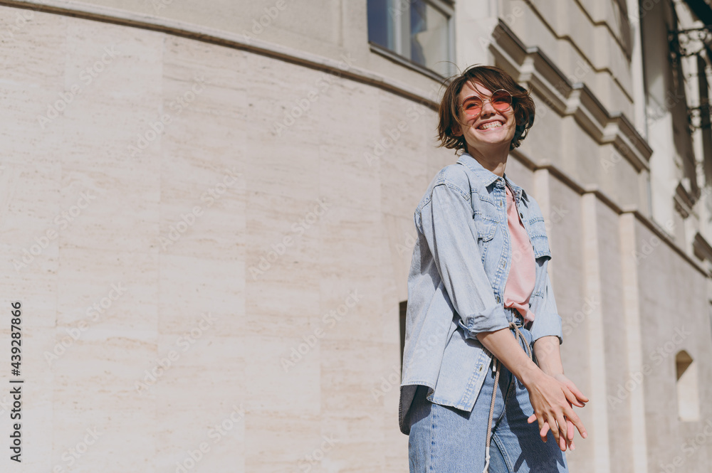 Young smiling fun happy cheerful stylish student caucasian woman 20s in jeans clothes eyeglasses walking going down street near building wall outdoors look aside People active urban lifestyle concept