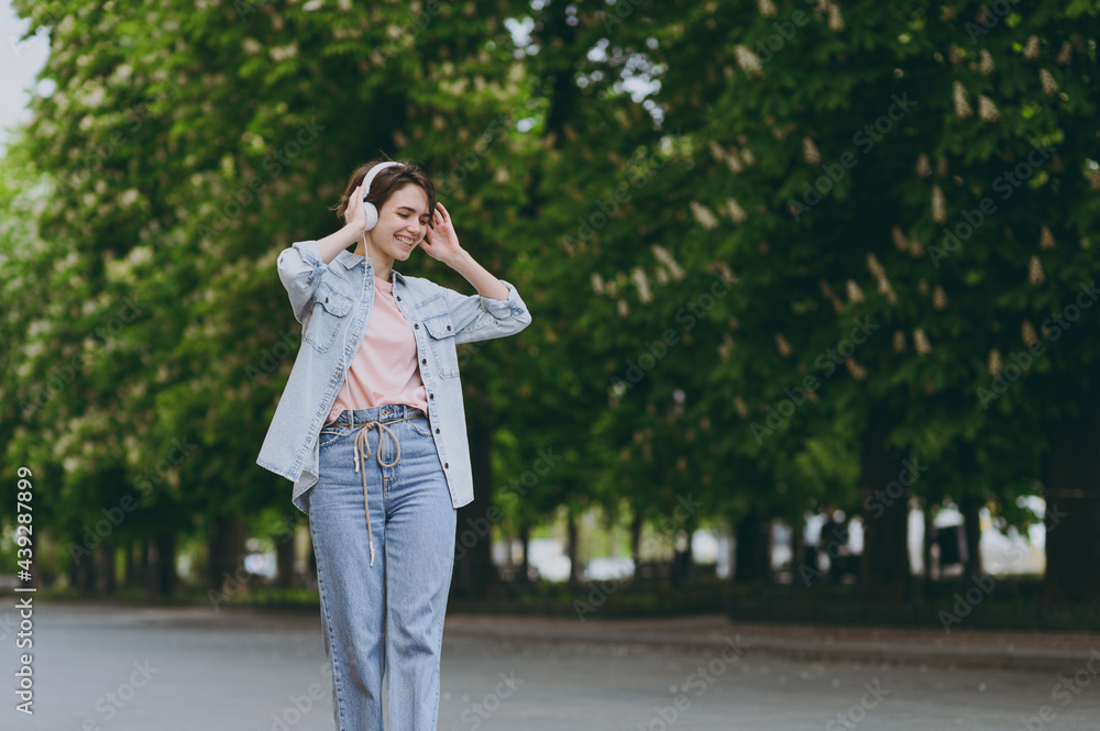 Young satisfied happy student stylish caucasian woman 20s wearing jeans clothes headphones listen to music walking strolling down green park alley outdoors strolling People urban lifestyle concept Young satisfied happy student stylish caucasian woman 20s wearing jeans clothes headphones listen to music walking strolling down green park alley outdoors strolling People urban lifestyle concept