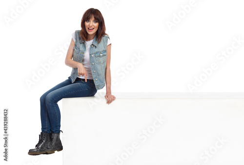 Ecstatic Young Woman Is Sitting On A White Box, Pointing Down And Talking