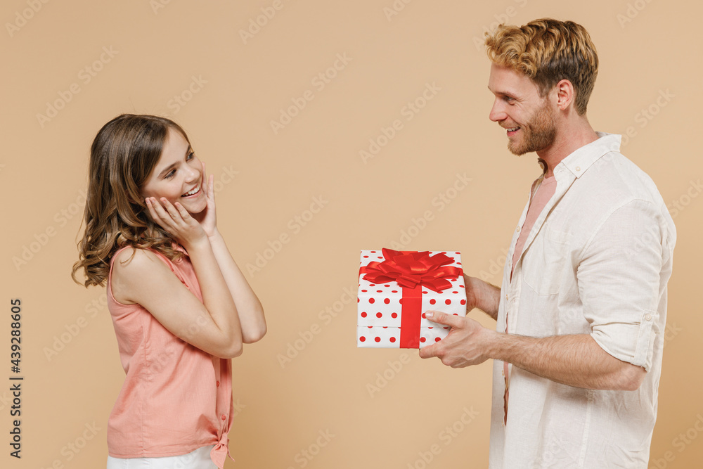 Side view young surprised parent man with child teen girl in casual clothes Dad little kid daughter red present box with gift ribbon bow isolated on beige background. Father's Day birthday concept Side view young surprised parent man with child teen girl in casual clothes Dad little kid daughter red present box with gift ribbon bow isolated on beige background. Father's Day birthday concept