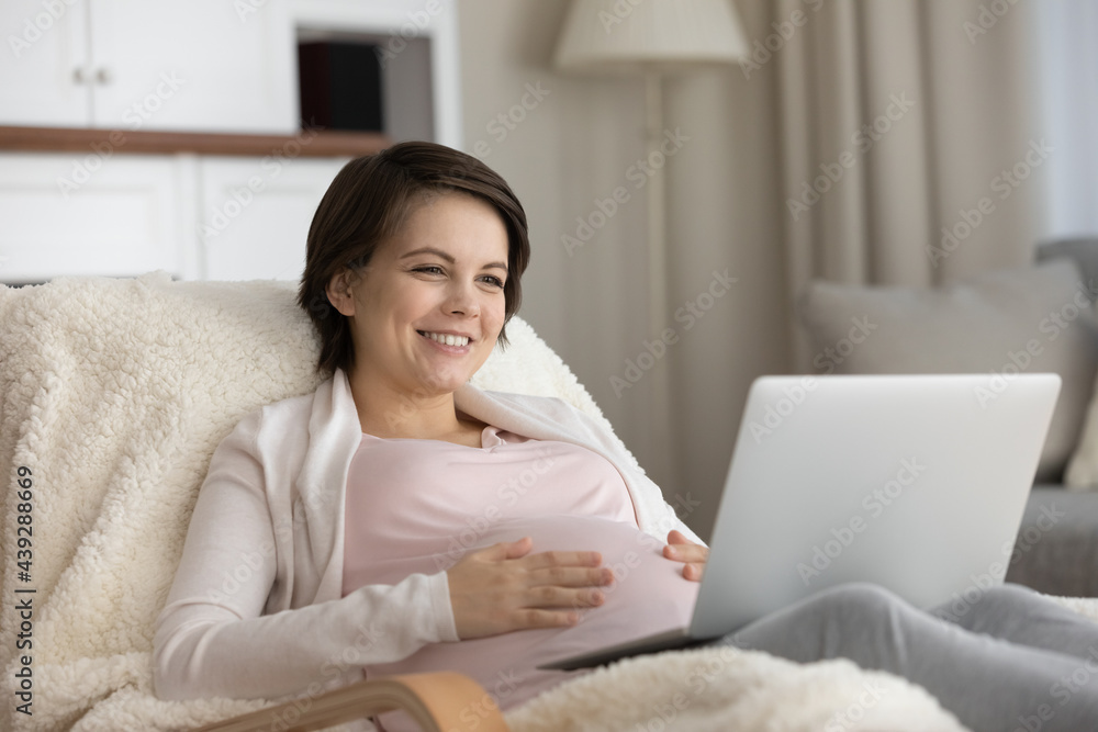 Smiling pregnant woman using laptop, looking at screen, touching caressing belly, sitting in cozy chair at home, happy young future mom watching movie or making video call, shopping online Smiling pregnant woman using laptop, looking at screen, touching caressing belly, sitting in cozy chair at home, happy young future mom watching movie or making video call, shopping online