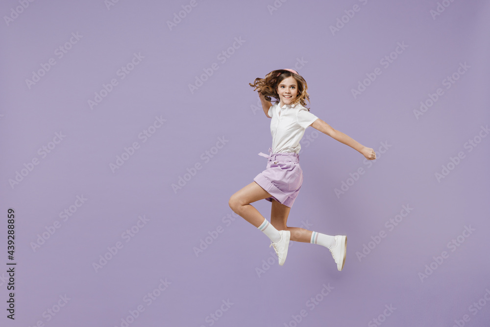Full length little fun kid girl 12-13 years old wear white short sleeve shirt jumping high with outstretched hands like fly isolated on purple color background. Childhood children lifestyle concept
