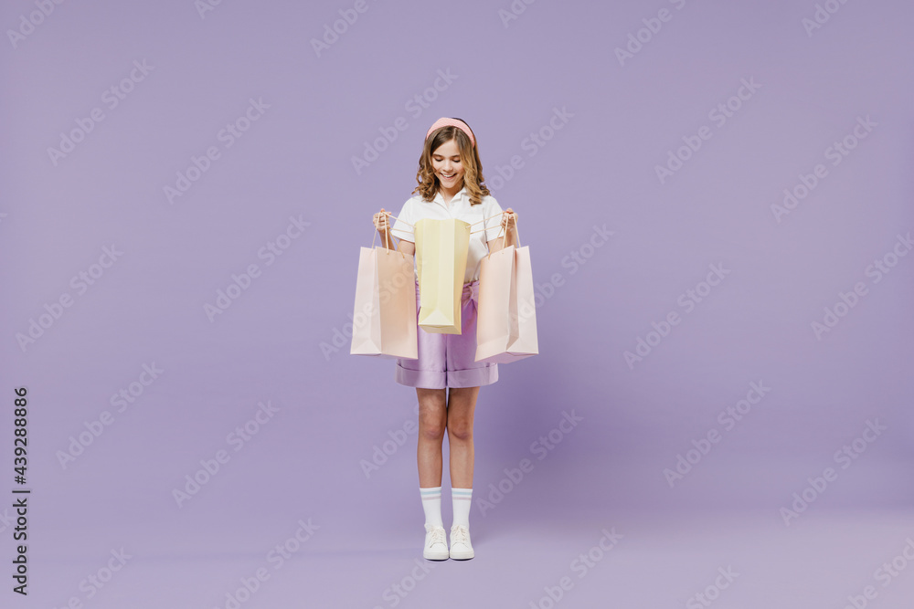 Full length fun smiling little kid girl 12-13 years old wear white shirt holding package bags with purchases after shopping isolated on purple color background. Childhood children lifestyle concept.