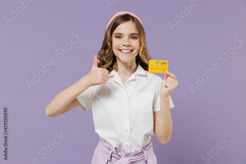 Little fun happy kid girl 12-13 years old in white short sleeve shirt holding credit bank card show thumb up gesture isolated on purple background children studio portrait Childhood lifestyle concept