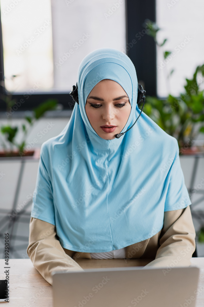 pretty muslim woman in headset working near blurred laptop in call center pretty muslim woman in headset working near blurred laptop in call center
