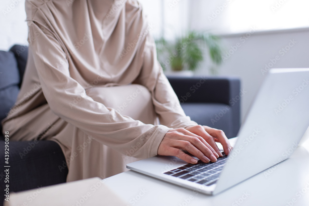 Fototapeta premium cropped view of muslim woman typing on laptop on blurred foreground