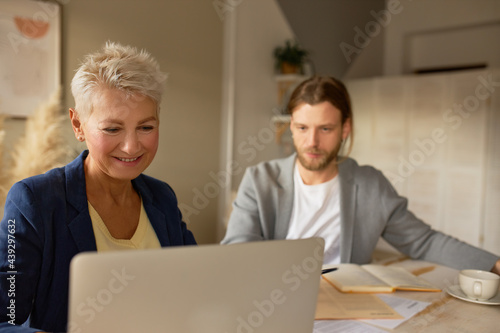 Business and cooperation concept. Successful middle aged female and bearded male manager working together at office, sitting in front of open laptop, having online business meeting using video chat