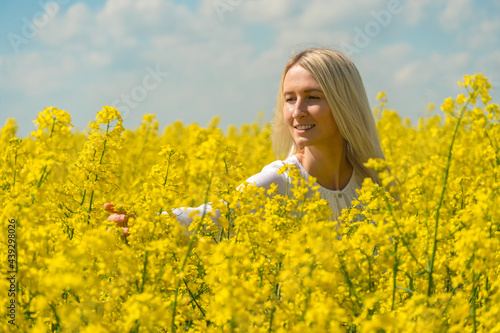 Smiling young blonde woman with her arm outstretched in a white shirt in a rapeseed field touching flowers on a sunny day