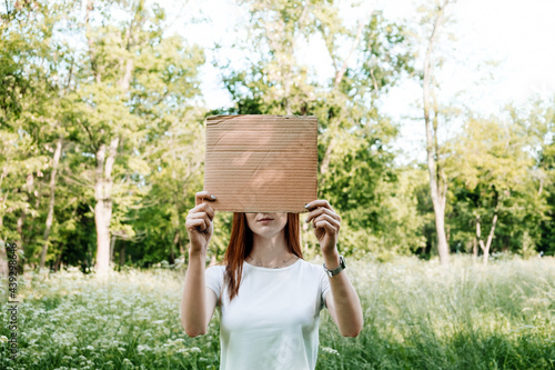 Outdoor mockup Young redhead woman covers her face with cardboard poster. Faceless mock up portrait of woman holding cardboard sign on nature background. © irissca