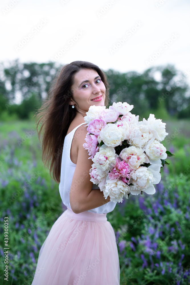 Fototapeta premium Beautiful girl with pink peonies, enjoying a bouquet of flowers on background of nature. Happy smiling woman in dress holds peonies in hands. Girl with a bouquet of flowers. Girl in wildflowers. 