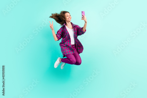 Full length body size view of trendy beautiful cheery girl jumping taking selfie showing v-sign isolated over bright teal color background