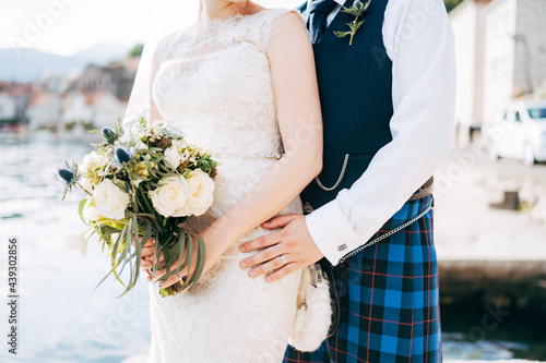 A bride in a wedding dress with bridal bouquet and a groom in a Scottish national dress stand hugging on the pier 