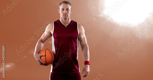 Portrait of caucasian male basketball player holding basketball against spot of light in background