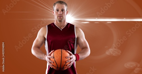 Portrait of caucasian male basketball player holding basketball against spot of light in background