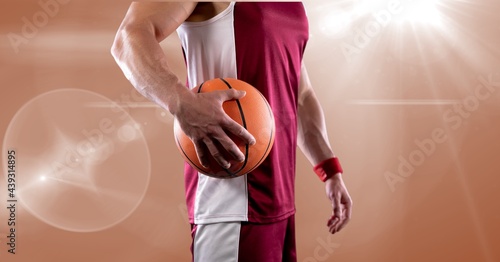 Mid section of male basketball player holding basketball against spot of light in background