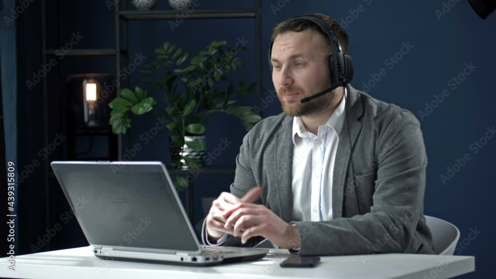 Young business man in a headset sitting at an office table in front of a laptop. A man is talking with a partner via a video call. Young business man in a headset sitting at an office table in front of a laptop. A man is talking with a partner via a video call.