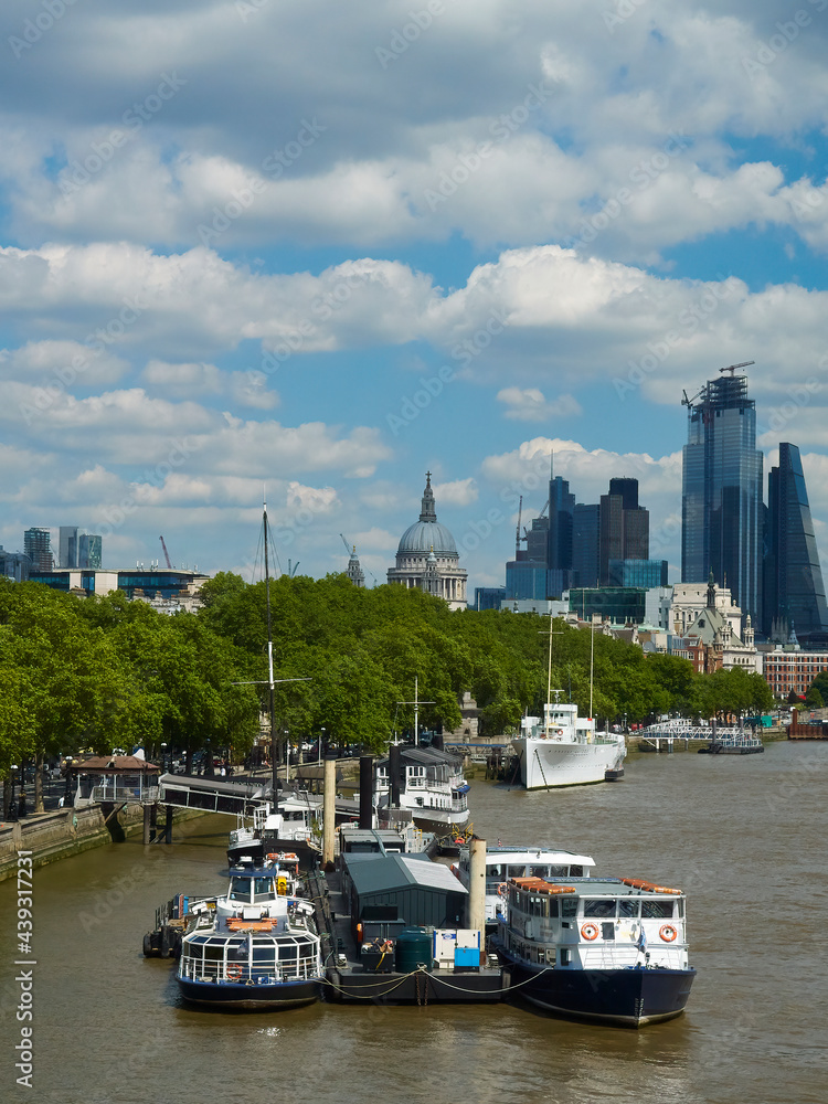 A view along the Victoria Embankment, passed a boat-filled jetty and riverside trees to the St. Paul’s Dome and the skyscrapers of the City of London.