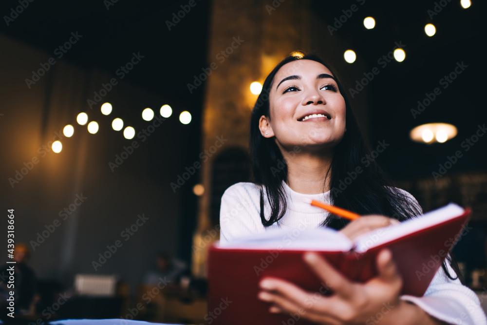 Happy Caucasian poet satisfied with creation process sitting at table with education textbook and smiling, cheerful female with brunette hair enjoying leisure time for planning week schedule - obrazy, fototapety, plakaty 