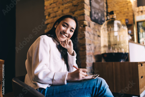 Content attractive Asian female student making notes in clipboard with sheet of paper and using wireless earphones while sitting in cafe