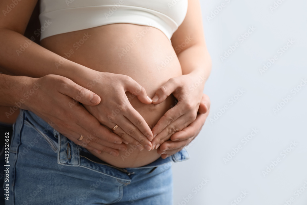 Pregnant young woman making heart with hands on belly and husband near her against light background, closeup Pregnant young woman making heart with hands on belly and husband near her against light background, closeup