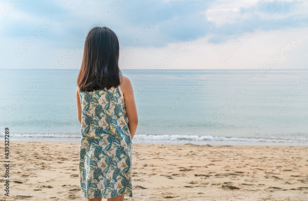 Asian child girl standing alone on the beach. Looking at the vast blue sky and the emerald sea. View from behind the girl, black long hair, empty space for text. Asian child girl standing alone on the beach. Looking at the vast blue sky and the emerald sea. View from behind the girl, black long hair, empty space for text.