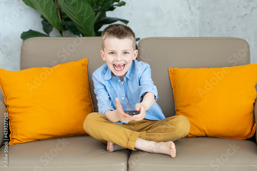 Smiling little boy watching TV and uses a tv remote sitting on sofa in the living-room