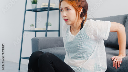 Young Korean lady in sportswear exercises doing working out doing tricep dips leaning on couch in living room at home. Social distance, Isolation during the virus. Exercises for the lower body.