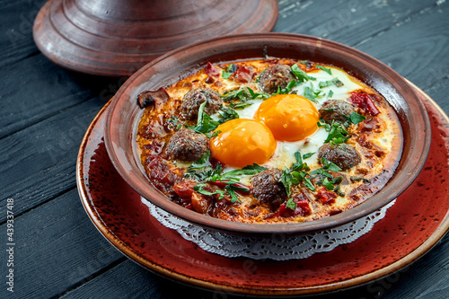 Classic Israeli scrambled eggs - shakshuka with tomatoes, herbs and meat kebabs, served in a clay plate on a black background.