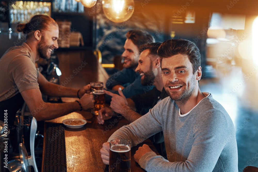 Smiling young men in casual clothing drinking beer and bonding together while sitting in the pub