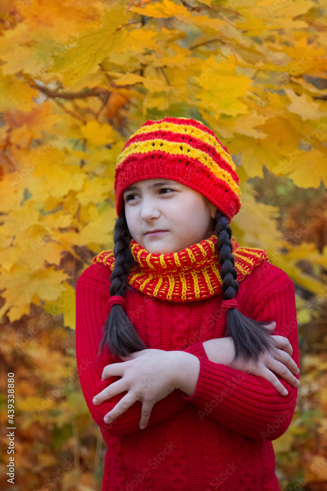 A girl in a red and yellow hat with dark hair looks offended and irritated to the side. On an autumn forest or park with yellow maple leaves A girl in a red and yellow hat with dark hair looks offended and irritated to the side. On an autumn forest or park with yellow maple leaves