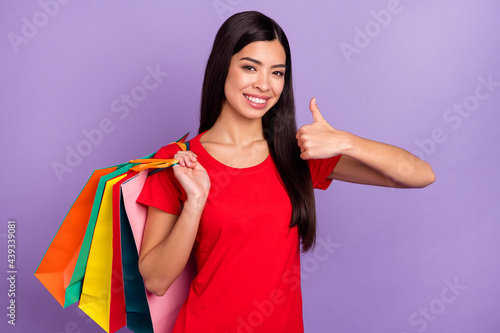 Photo of sweet confident woman dressed red t-shirt holding back colorful bargains showing thumb up isolated purple color background