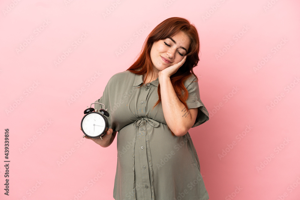 Fototapeta premium Young redhead caucasian woman isolated on pink background pregnant and holding clock doing sleep gesture