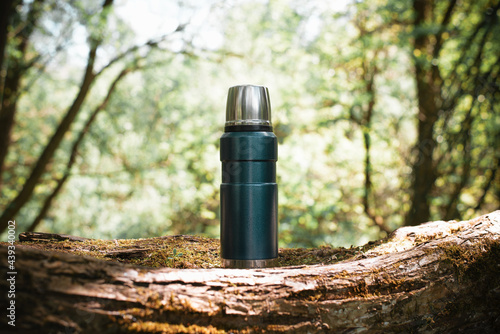 Thermos standing on a tree outdoors, blurred backdrop forest sunny summer day. Hiking flask with a hot drink in nature. Camping concept.