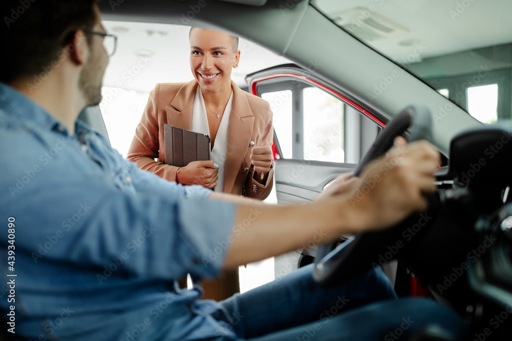Saleswoman is showing new car to young Caucasian man in showroom. Saleswoman is showing new car to young Caucasian man in showroom.