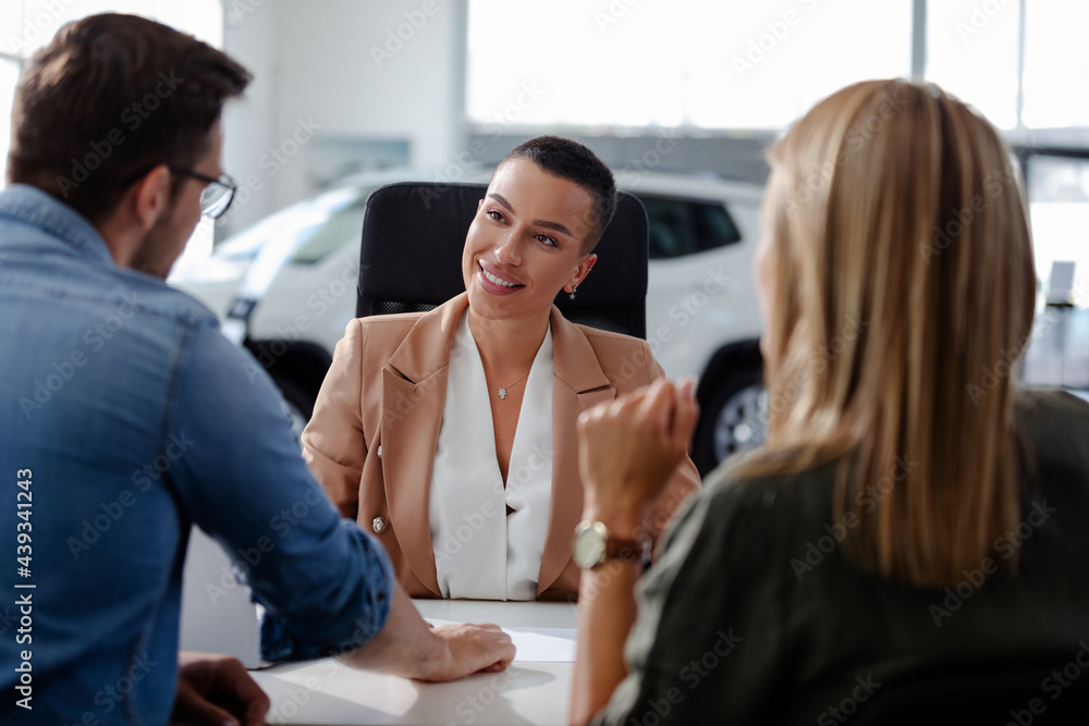 Smiling saleswoman talking with young couple about contract for new car in modern car showroom.