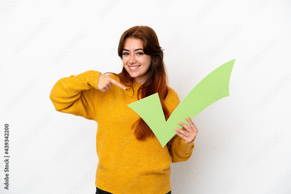 Young redhead woman isolated on white background holding a check icon and pointing it Young redhead woman isolated on white background holding a check icon and pointing it
