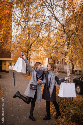 Two handsome young women are standing in the park, posing with bags in their hands and looking at each other. Autumn and shopping