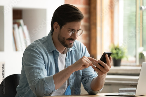 Millennial generation businessman in glasses using cellphone software app, checking emails, communicating distantly with clients or relaxing watching funny video photo content, enjoying break time.