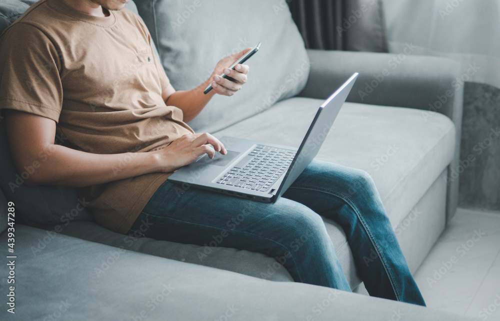 Naklejka premium young man typing on mobile phone while sitting on a sofa at home with laptop computer. Man using his smartphone for online banking.work from home concept.