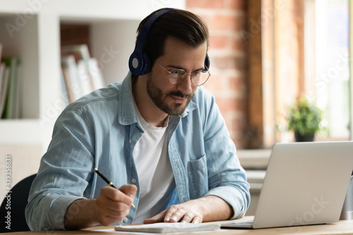 Happy focused young man in eyeglasses wearing wireless headphones, watching educational lecture on computer, studying on online courses distantly, improving professional knowledge, writing notes.