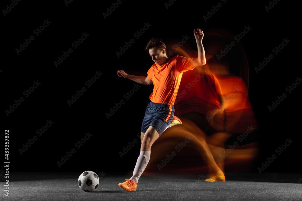 Young caucasian male football or soccer player kicking ball for the goal in mixed light on dark background. Concept of healthy lifestyle, professional sport, hobby.