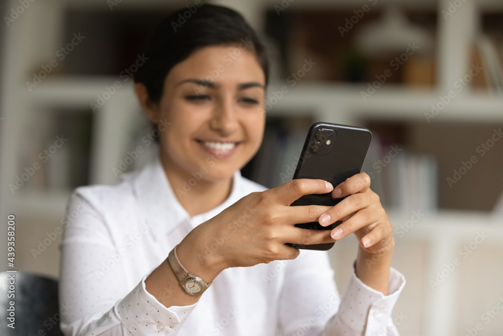Happy young indian multiracial woman holding smartphone in hands, using mobile software application, web surfing information online, communicating distantly in corporate social network in office. Happy young indian multiracial woman holding smartphone in hands, using mobile software application, web surfing information online, communicating distantly in corporate social network in office.