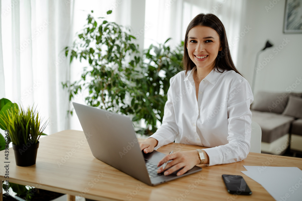 Photo of cheerful pretty student woman smiling and using laptop while doing homework in living room. Photo of cheerful pretty student woman smiling and using laptop while doing homework in living room.