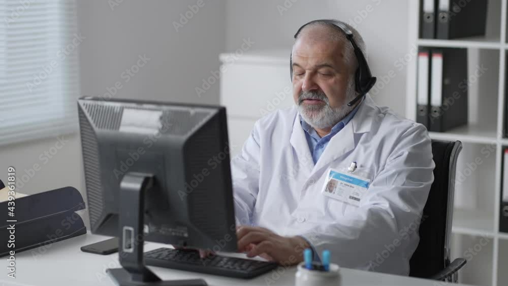 medicine, technology and healthcare concept - senior male doctor or nurse with headset and computer working at hospital. Ambulance support hotline operator medicine, technology and healthcare concept - senior male doctor or nurse with headset and computer working at hospital. Ambulance support hotline operator