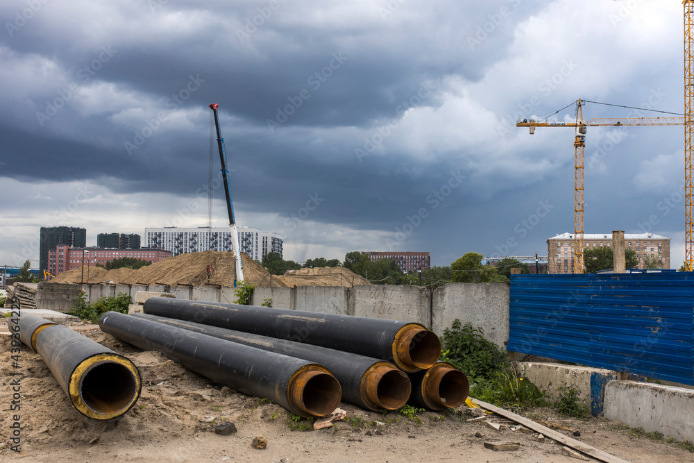 Long factory pipes lie on the ground against the backdrop of a residential area under construction Long factory pipes lie on the ground against the backdrop of a residential area under construction
