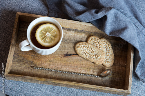 A white cup of tea with lemon, a long cupronickel spoon with a twisted handle and a saucer with three oatmeal cookies for breakfast on a wooden tray, a rag napkin on a gray sofa. photo